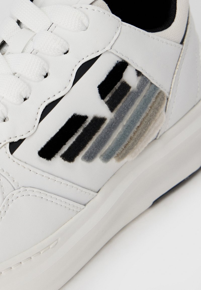 Sneaker bianco con suola in gomma testurizzata, presenta strisce in peluche nere, grigie e blu sul lato. I lacci e le cuciture sono bianchi.