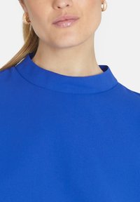 Kobaltblå topp med hög, rundad halsringning och mjuk textur. Guldtonade, cirkulära örhängen kompletterar outfiten.