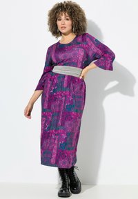 Lila gemustertes Midi-Kleid mit weiten Ärmeln und einem taillierten grauen Gürtel, kombiniert mit schwarzen Schnürstiefeln. Glatter Stoff mit aufwendigen Mustern.