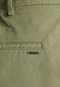 Gros plan d'un pantalon en tissu vert montrant une poche arrière cousue avec une petite étiquette de marque en métal "HUGO" sous la poche.