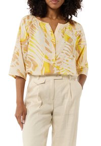 Kurzarmbluse mit einem gelb-beigen tropischen Muster, V-Ausschnitt und lässigem Schnitt, kombiniert mit hellbeigen, hochgeschnittenen Hosen.