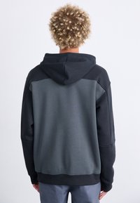 Sudadera negra y gris con un corte holgado, que presenta una capucha grande y un diseño bicolor con texturas contrastantes en la parte posterior.