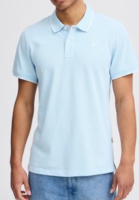 Svijetloplava polo majica s ovratnikom i bijelim detaljima, koja ima kratke rukave, kopčanje na tri gumba i teksturiranu tkaninu.
