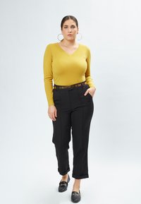 Gelber V-Ausschnitt-Pullover, schwarze maßgeschneiderte Hose mit umgeschlagenen Säumen und schwarze Slipper. Dazu große Creolen und ein metallischer Gürtel.