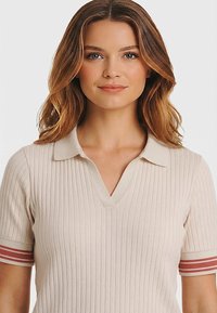 Joven con cabello castaño claro y ondulado que lleva una camiseta tipo polo de manga corta de color crema con puños a rayas rojas, sonriendo suavemente.