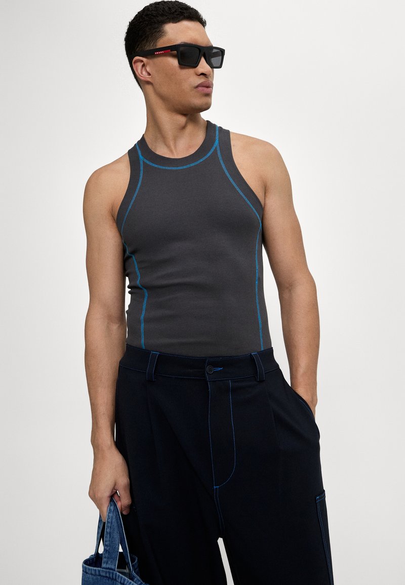 Graues Tank-Top mit blauen Akzenten, kombiniert mit navyfarbenen Hosen mit sichtbaren Nähten, gehalten mit einer Denim-Tote-Tasche und schwarzen Sonnenbrillen.