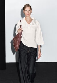 Bílá košile s širokým výstřihem, doplněná volnými tmavými jeansy. V pravé ruce drží texturovanou, tmavě červenou tkanou tašku.