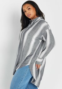 Camisa de botones a rayas grises y blancas con cuello, mangas largas y aberturas laterales, hecha de una tela ligera. Combinada con vaqueros azules.