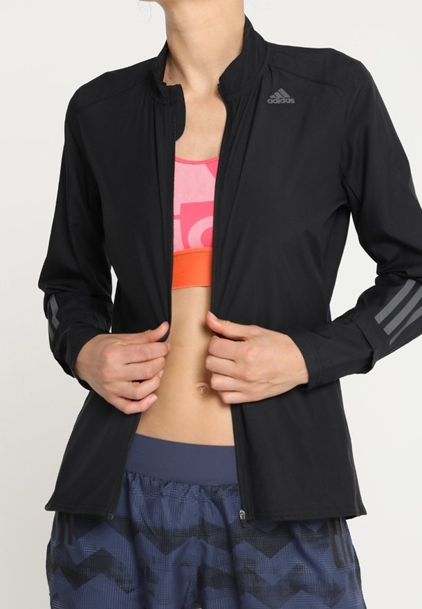Veste de sport noire avec col montant, fermeture éclair à l'avant, accent à trois bandes sur les manches, tissu léger. Portée par-dessus un haut de sport coloré.