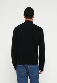 Fekete kötött pulóver magas gallérral és kényelmes szabással. Az anyag puhanak tűnik, hosszú ujjakkal és sima textúrával rendelkezik.