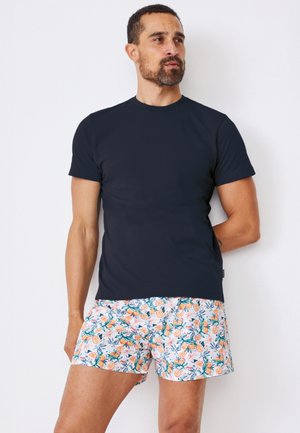 Mężczyzna ubrany w gładką czarną koszulkę i białe szorty z pomarańczowo-zielonymi wzorami owoców i liści, stojący na białym tle.