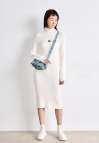 Vestido de punto blanco acanalado con cuello alto, mangas largas y una abertura lateral. Combinado con un bolso cruzado azul claro y zapatillas blancas.