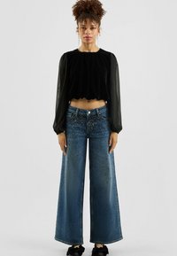 Zwarte cropped top van fluweel met geplooide details, gecombineerd met wijde blauwe denim jeans met gespen aan de voorkant.