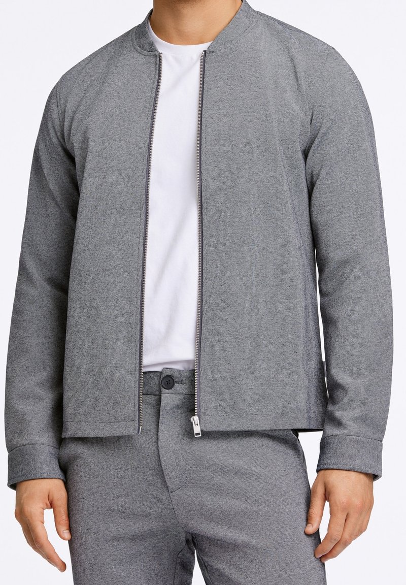 Homme portant une veste zippée gris clair sur un T-shirt blanc et un pantalon gris assorti, debout les bras détendus le long du corps.