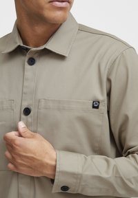 Beige shirt with a buttoned collar, two front pockets, and black button accents. Smooth texture with a logo on the chest pocket.