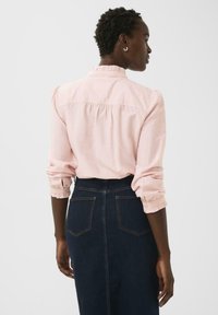 Femeie cu păr scurt și creț, purtând o bluză cu mânecă lungă de culoare roz pal, băgată într-o fustă din denim albastru închis cu talie înaltă, întoarsă cu spatele.