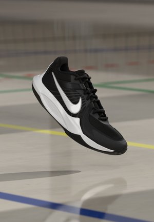 Chaussure de sport Nike noire et blanche suspendue au-dessus d'un terrain de sport couvert avec des lignes colorées.