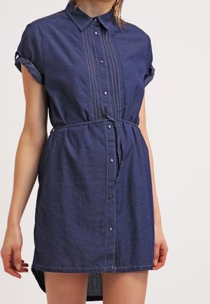 Kobieta ubrana w granatową sukienkę na guziki z krótkim rękawem z podwiniętymi rękawami i cienkim paskiem w talii, stojąca na jednolitym tle.