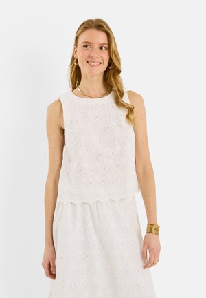 Femme aux longs cheveux blonds sourit, portant un top blanc sans manches à motifs œillets et une jupe assortie, des boucles d'oreilles anneaux en or, un collier et un bracelet.