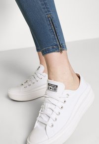 Zapatillas de lona blancas con suelas de goma, combinadas con jeans skinny azules que tienen un detalle de cremallera lateral y costuras en contraste.
