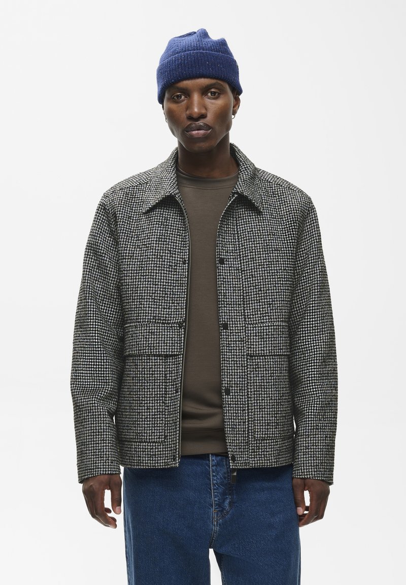 Homme portant un bonnet en tricot bleu, une veste pied-de-poule noire et blanche, une chemise marron et un jean bleu, debout devant un fond uni.
