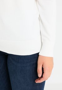 Valkoinen neulepaita, jossa on resorit hihansuissa, yhdistetty tummansinisiin farkkuihin. Keskitytään hihojen yksityiskohtiin ja materiaalin tekstuuriin.