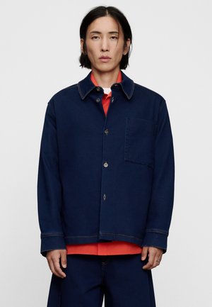 Person mit mittellangen schwarzen Haaren, die eine dunkelblaue, geknöpfte Jacke über einem roten Hemd trägt und vor einem einfarbigen Hintergrund steht.