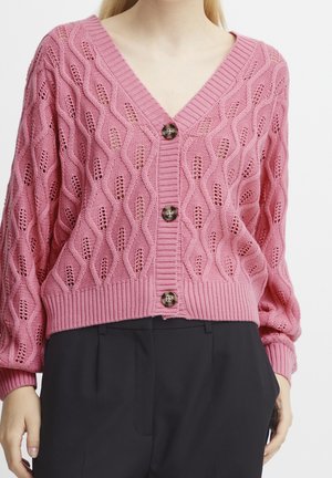 Kobieta w różowym dzianym kardiganie z wzorem warkoczowym i ażurowym, z brązowymi guzikami, w zestawieniu z czarnymi spodniami na białym tle.