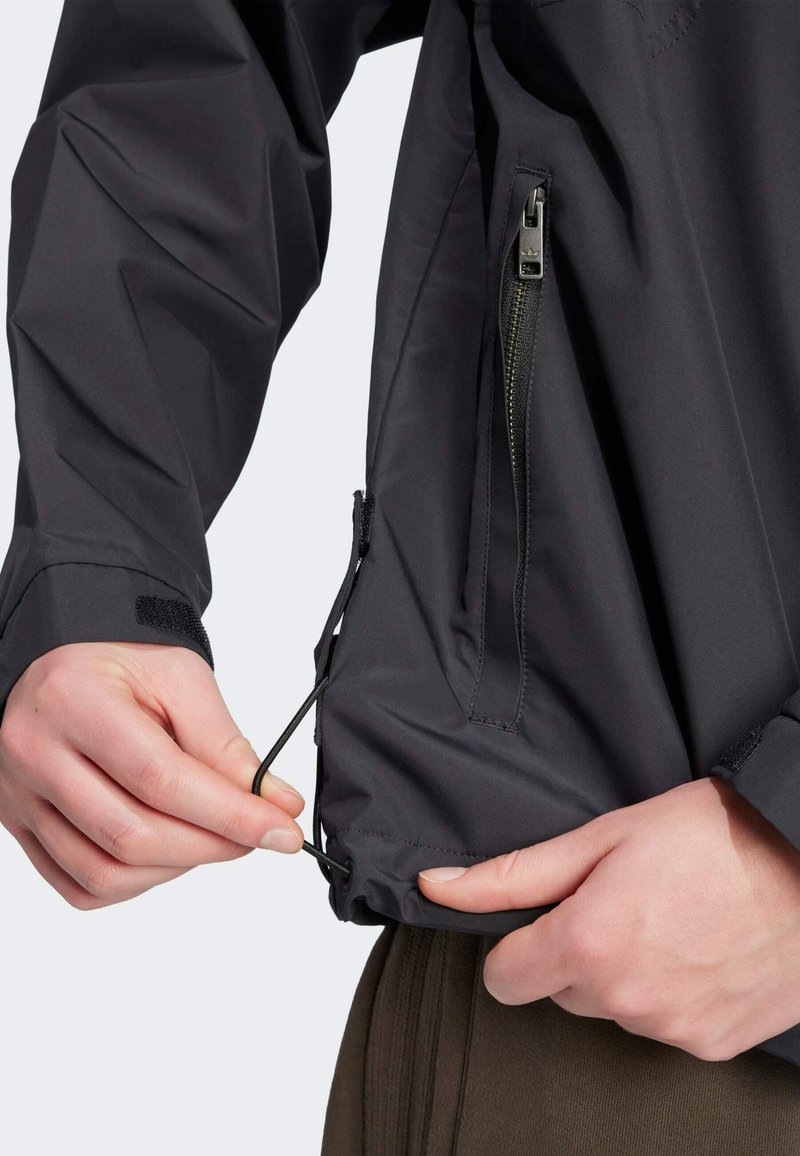 Veste imperméable noire avec un ourlet à cordon de serrage, poches latérales zippées et ajusteurs de poignet en Velcro, mettant en avant une texture lisse et mate.