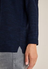 Granatowy sweter dzianinowy z teksturowanej tkaniny, z bocznymi rozcięciami na dolnym brzegu i prążkowanym okrągłym dekoltem. W połączeniu z jasnoszarymi dżinsami.