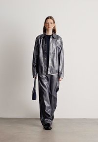 Casaco prateado metálico com fecho de botões na frente, combinado com calças largas a condizer. O modelo segura uma pequena mala preta. Textura suave e corte descontraído.