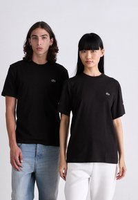 Dva modelky v černých tričkách s krátkým rukávem Lacoste, jedno v kombinaci s světle modrými džíny a druhé s bílými kalhotami, na jednolitém pozadí.