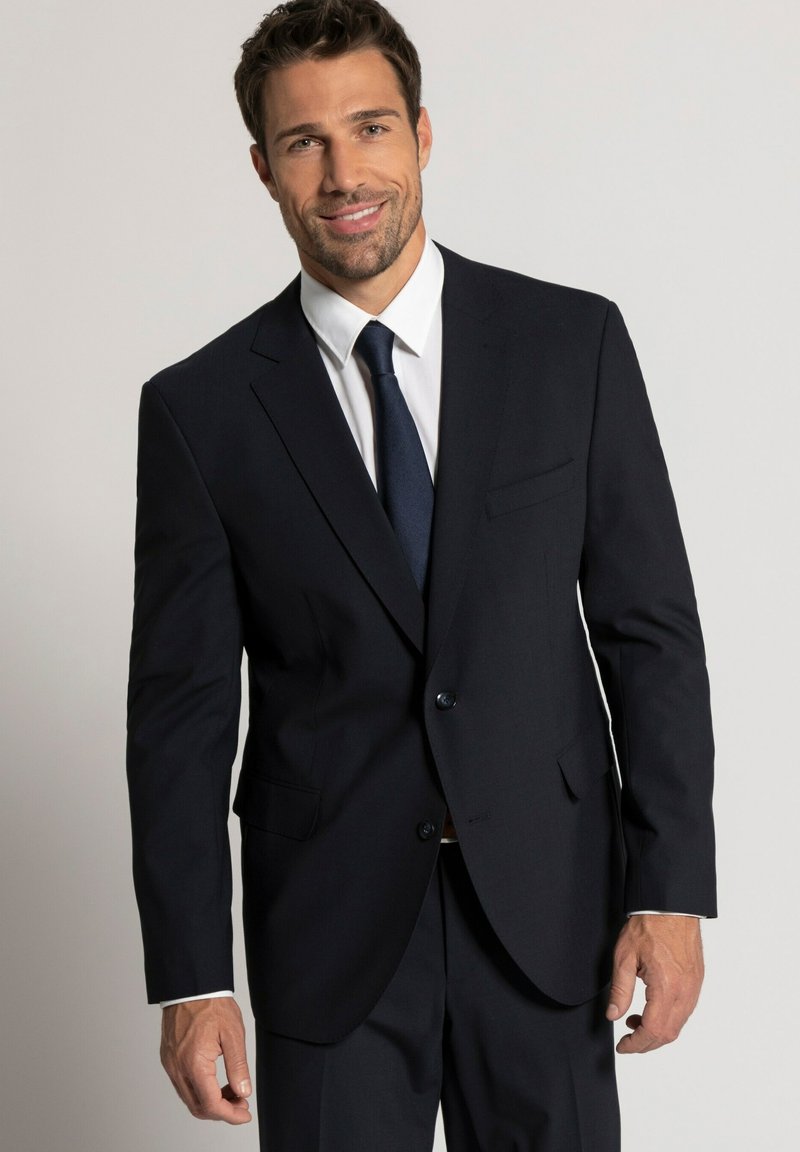 Giacca da abito blu navy con rever a pattina, chiusura a un bottone, due tasche frontali, abbinata a una camicia bianca e una cravatta scura. Texture liscia.