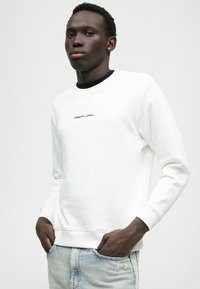 Balts sporta krekls ar melnu apkakli, centrā ar nelielu "REPLAY" logotipu. Audums ir mīksts un gluds, ar ribotām piedurknēm.