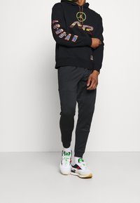 Sudadera con capucha negra con gráfico multicolor de "Jordan" en la manga, combinada con pantalones jogger oscuros y zapatillas deportivas blancas con detalles.