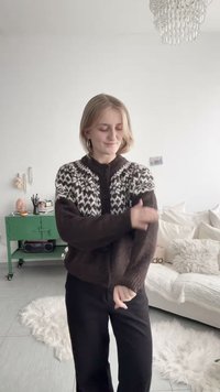 Joven ajusta la manga de un suéter de punto con estampado marrón y blanco en una sala de estar luminosa con cojines mullidos y un carrito verde.