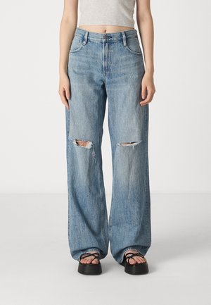 Osoba ubrana w jasnoniebieskie dżinsy o szerokich nogawkach z podartymi kolanami, czarne sandały na platformie oraz szary krótki top na tle jednolitego tła.