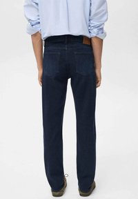 Persona con jeans azul oscuro y una camisa clara de manga larga arremangada, de pie con las manos relajadas a los lados, de espaldas a la cámara.