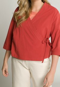 Rotes Wickeloberteil mit dreiviertel Ärmeln, seitlicher Bindung, strukturiertem Stoff und lockerem Schnitt, kombiniert mit hellbeigen Hosen.