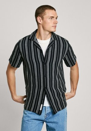 Camisa negra de manga corta con rayas verticales blancas, con botones en el frente y un cuello relajado, llevada sobre una camiseta blanca. Jeans de mezclilla visibles.