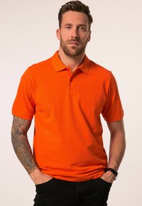 T-Shirt mit kurzen Ärmeln in leuchtendem Orange, aus Baumwolle, mit einem Kragen und einer Knopfleiste mit drei Knöpfen. Das Modell trägt schwarze Hosen.