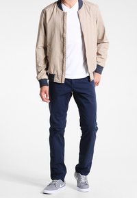 Man wearing beige bomber jacket with navy cuffs, white t-shirt, navy pants, and gray sneakers standing against white background.