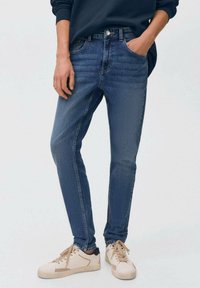 Slim-fit temno modri kavbojki z zbledelim teksturo, petimi žepi in gumbom za zapiranje, v kombinaciji z svetlimi supergami.