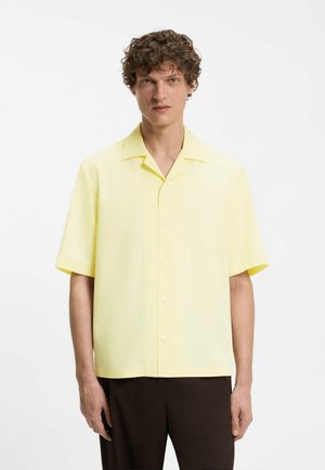 Ung mand med krøllet hår iført en kortærmet, lys gul skjorte med knapper og mørke bukser, stående mod en ensfarvet hvid baggrund.