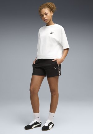 Femme portant un t-shirt Puma blanc, un short Puma noir, des chaussettes Puma blanches et des baskets Puma noires et blanches, debout les mains dans les poches.