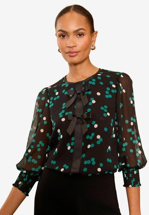 Schwarze Bluse mit türkisfarbenen und weißen Punkten, ausgestattet mit Puffärmeln und zwei Stoffschleifen auf der Vorderseite. Durchsichtiges Material, geraffte Bündchen.