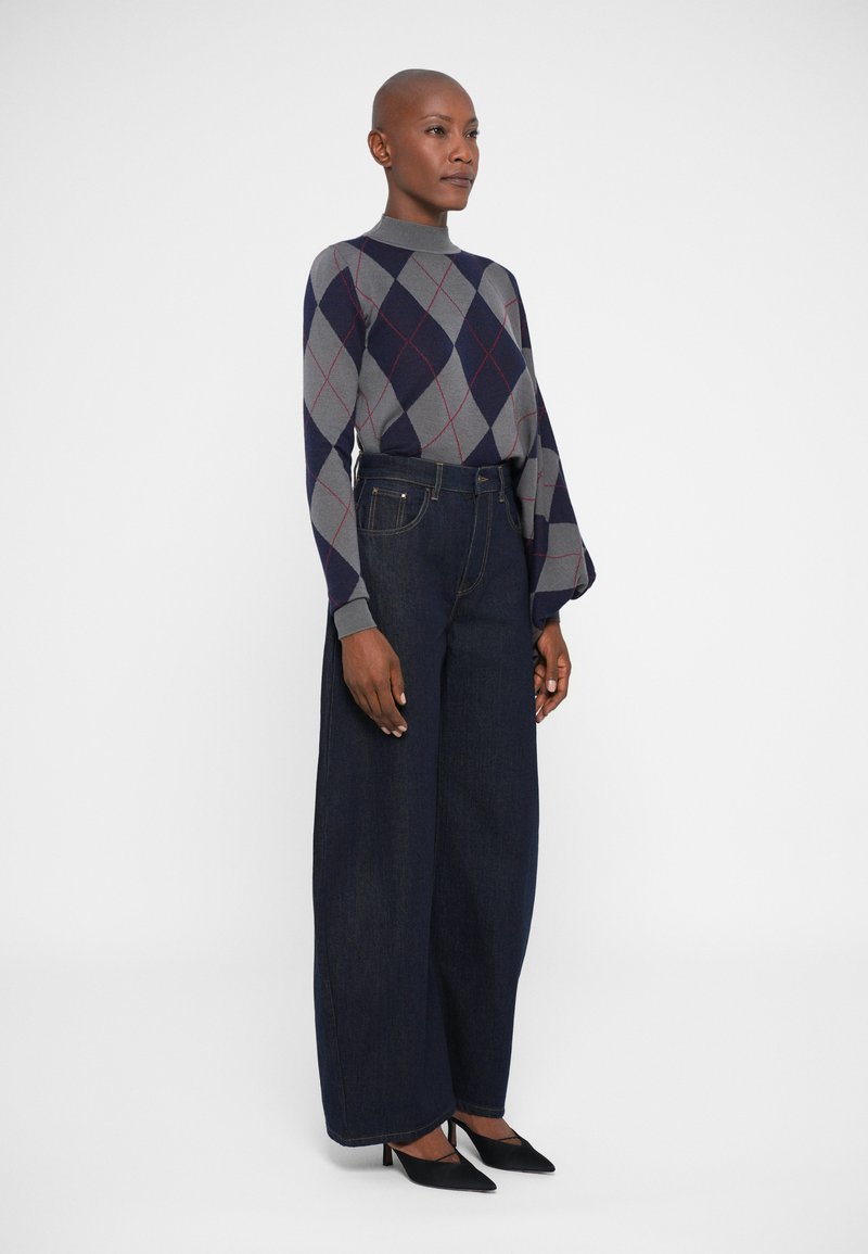 Maglione argyle grigio e blu navy con un alto collo, abbinato a jeans scuri a gamba larga e décolleté nere a punta.