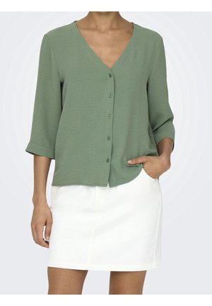 Vrouw die een saliegroene blouse met knopen en driekwartmouwen draagt en een witte knie-lange rok, hand in de zak.