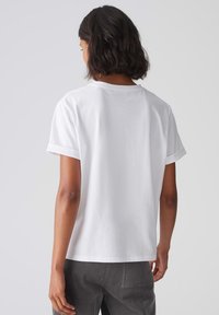 T-shirt bianco a maniche corte con risvolti, colletto rotondo e texture in cotone liscia, indossato con pantaloni in velluto a coste grigi, visto da dietro.
