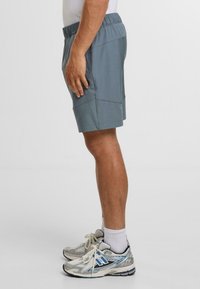 Šedé sportovní šortky s elastickým pasem, bočními kapsami a zesíleným šitím, doplněné šedými a modrými běžeckými botami.